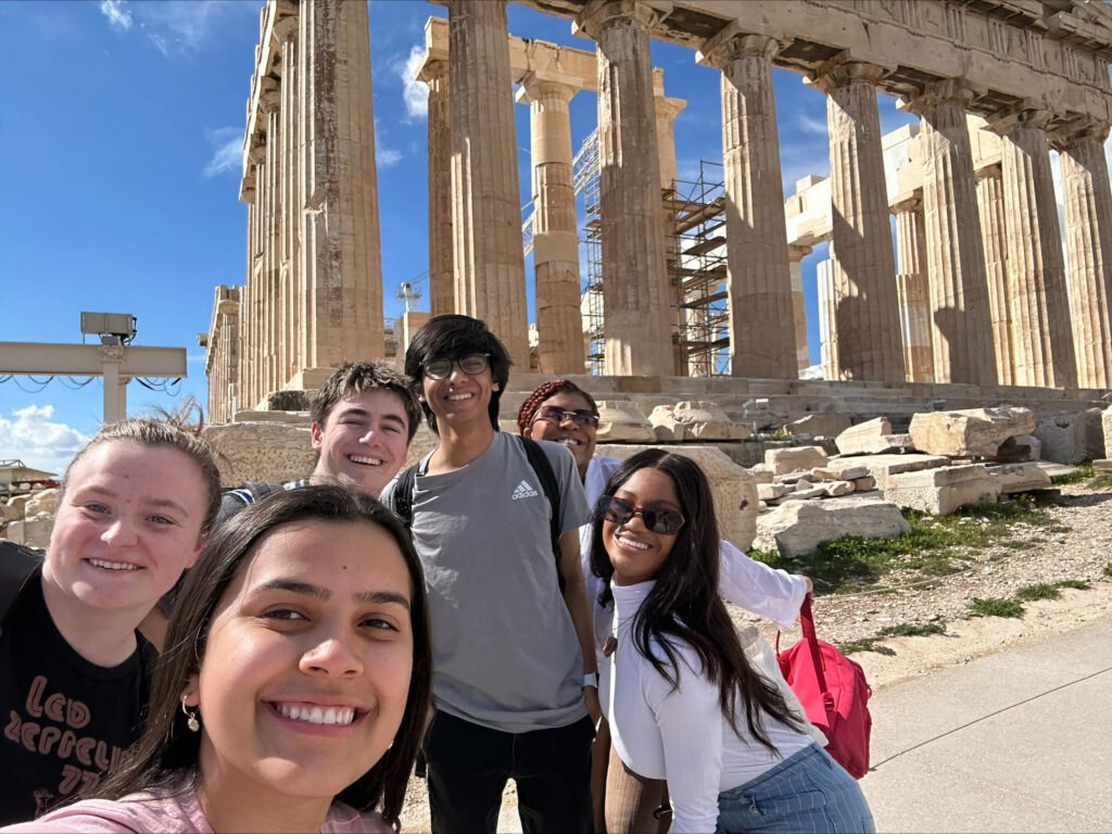 Group photo with Michelle Kengkart (center) and, left to right, Grace Pilch, Owen Perry, Mihir Kulkarni, Leila Filien, De'jah Coates, posing in front of the Parthenon in Athens, Greece.