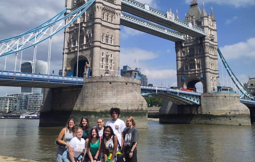 A group of Penn State students on the Literary London faculty-led course trip standing with the London Bridge in the behind them in the background.