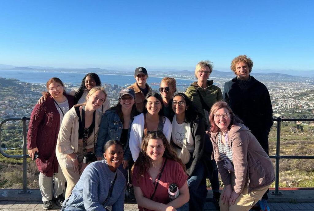 Students stand on top of a mountain with Cape Town, South Africa, in the distance behind them. From left to right, back row: Monica Rodriguez, Safian Lewis, Corinne Chase, Jill Wood, Susanne M. Klausen, Ethan Capitano. Middle row: Payton Smith, Cara Arnoldi, Samantha Powell, Keya Ahrestani, Hannah Yurack. Front row: Phathutshedzo Bale (local staff member), Maggie Day