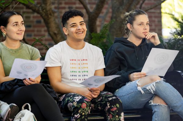 Three students in a Spanish class sit on a bench outside Willard Building on a nice day.