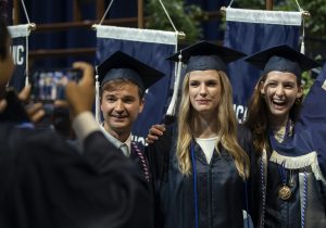 Three students dressed in academic regalia smile at someone taking their photo at the Liberal Arts commencement ceremony.