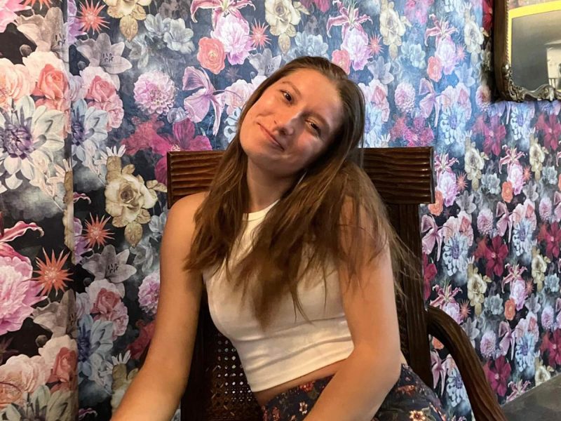 Anna Raquel Duiker, who will graduate Penn State in 2026, smiles while seated, with a flowery wallpaper in the background.