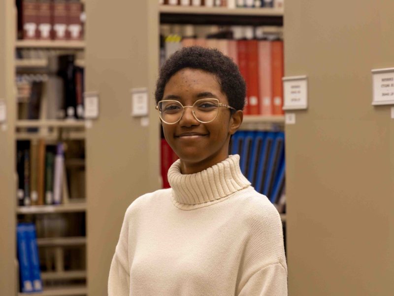 Jasmine Upchurch, a 2025 Penn State graduate, smiles while wearing glasses and a white sweater, in a library.