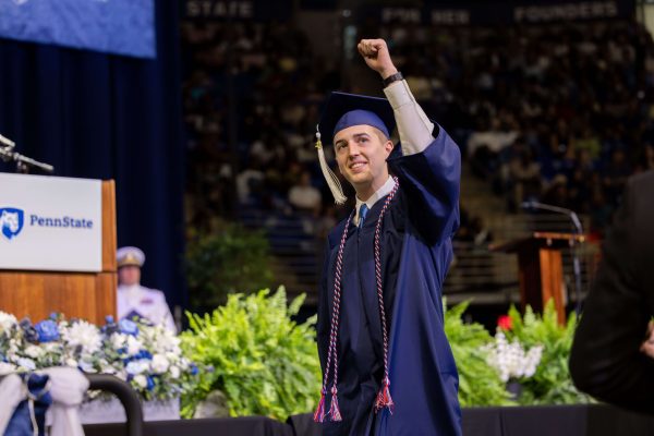 A Penn State graduate in a blue cap and gown raises a triumphant fist while walking across the commencement stage.