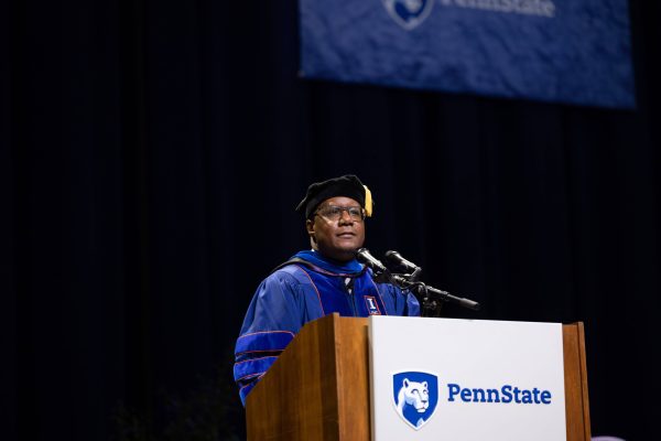 The Penn State College of Liberal Arts Dean, dressed in academic regalia with a gold tassel and blue robe, stands at a podium delivering a speech.