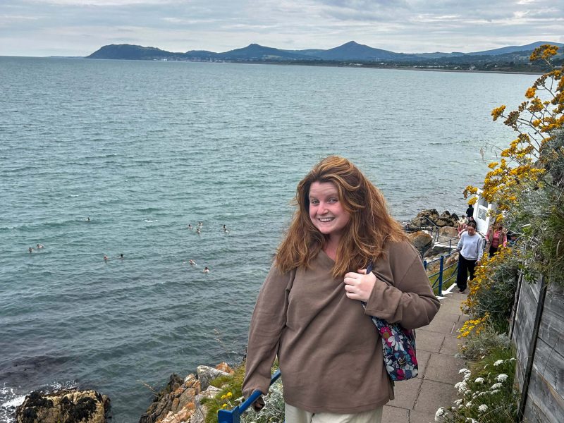 Teagan Mayr, a Perreault Fellow, standing on a cliffside walking path overlooking the water.