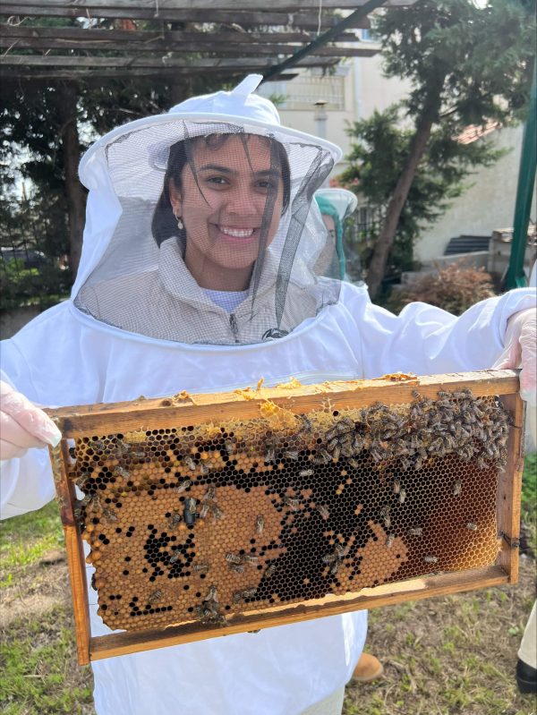 Michelle Kengkart wearing a beekeeper's white suit, holding a honeycomb panel while visiting From A to Bee Farm in Pallini, Greece.