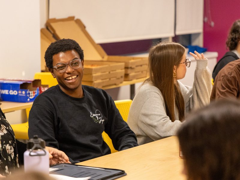 Zion Sykes, 2024–25 University Park Undergraduate Association president, sits in a chair at a table full of first-year Liberal Arts students while laughing and talking to the group.
