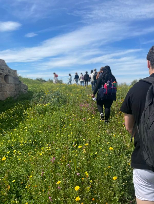 Group of Penn State students walking, with their backs facing the camera, through the fields of architectural remains at Aegina Island, Kolona.